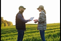 Caucasian man farmer standing in field with tablet device in hands and showing something to woman on screen.